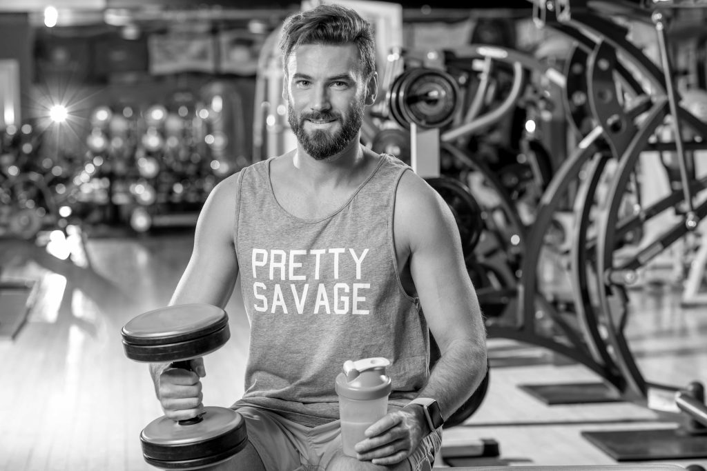 Hombre en gimnasio sosteniendo una mancuerna y un bote de mezcla de liquido, en el fondo maquinas de gimnasio, la foto esta en blanco y negro